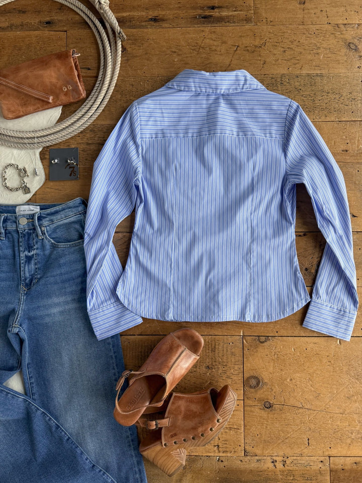 The Mandeville Blue Striped Pleat Top by Savannah Sevens western life{&}style, blue jeans, brown heeled sandals, a brown wallet, jewelry, and a coiled rope are arranged on a wooden floor.