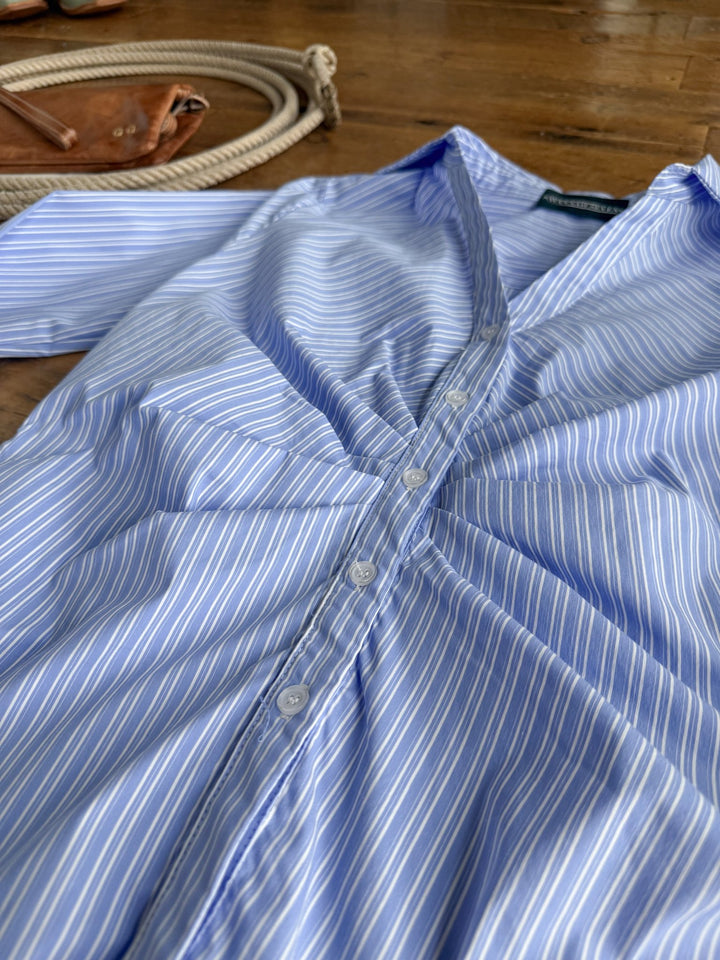 The Mandeville Blue Striped Pleat Top by Savannah Sevens western life{&}style is laid out on a wooden floor, with a brown leather bag and a coiled rope visible in the background.