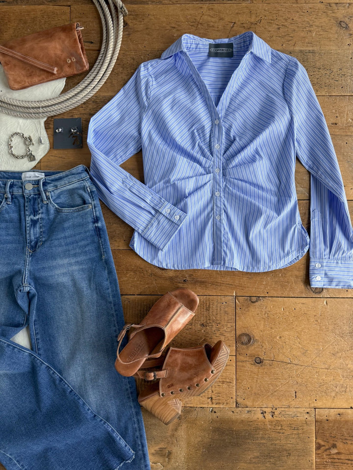A flat lay on a wood floor featuring the Savannah Sevens western life{&}style Mandeville Blue Striped Pleat Top, blue jeans, brown leather clogs, a small brown handbag, silver jewelry, and a coiled rope.