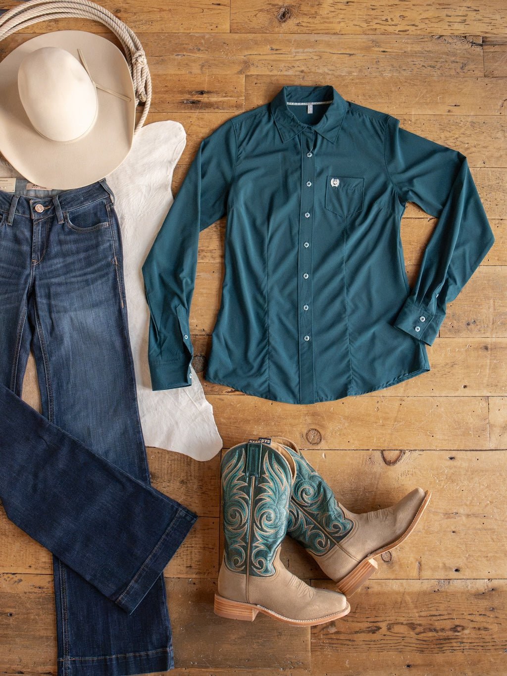 Flat lay of the Mankato Teal Mesh Performance Shirt from Savannah Sevens western life{&}style, paired with blue jeans, cream cowboy boots featuring teal embroidery, a white cowboy hat, and a white scarf arranged on a wooden floor.