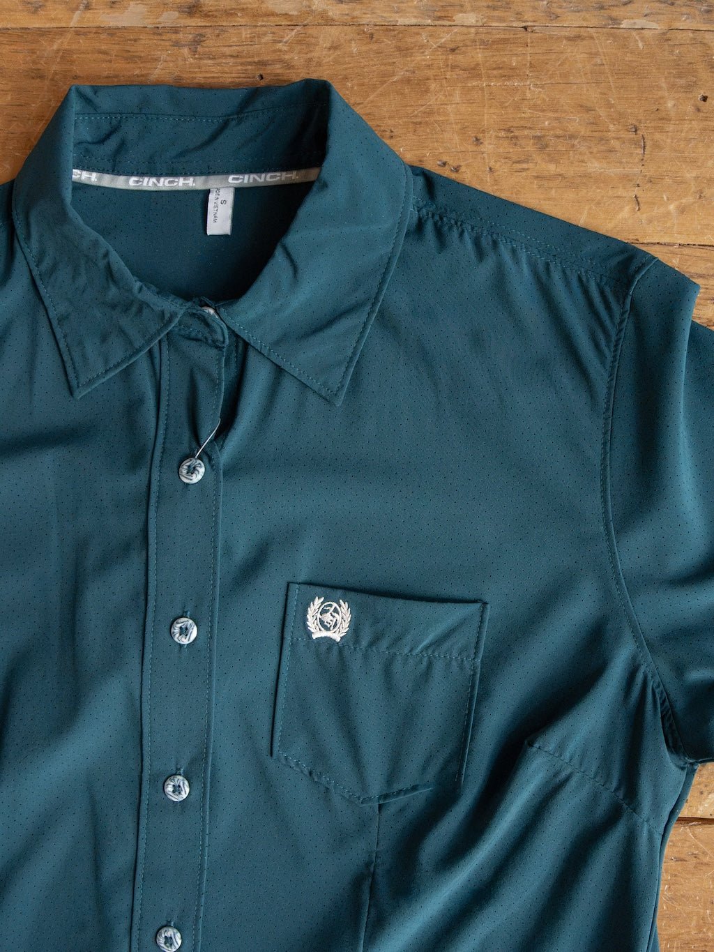 The Mankato Teal Mesh Performance Shirt by Savannah Sevens western life{&}style is a teal long sleeve button up with a chest pocket and a small white embroidered logo above the pocket. Laid out on a wooden surface, this quick-drying shirt provides added UV defense for extra comfort and protection.