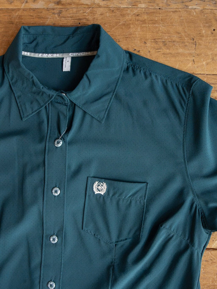 The Mankato Teal Mesh Performance Shirt by Savannah Sevens western life{&}style is a teal long sleeve button up with a chest pocket and a small white embroidered logo above the pocket. Laid out on a wooden surface, this quick-drying shirt provides added UV defense for extra comfort and protection.