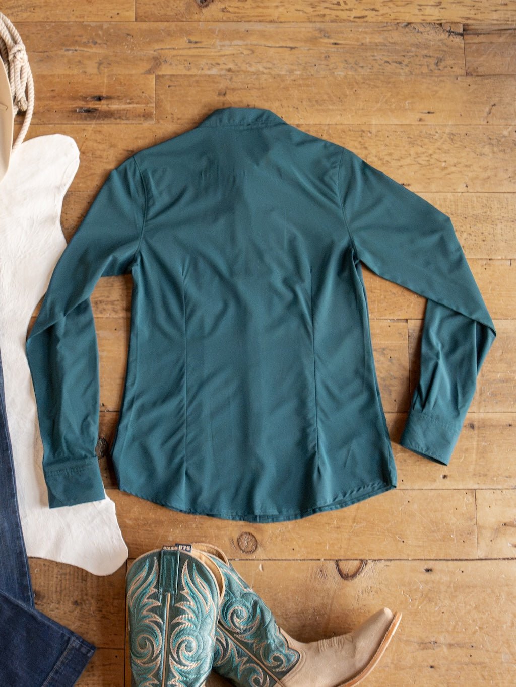 The Mankato Teal Mesh Performance Shirt by Savannah Sevens western life{&}style is laid flat on a wooden floor, accompanied by turquoise cowboy boots, with part of a white cowhide and denim jeans visible nearby.