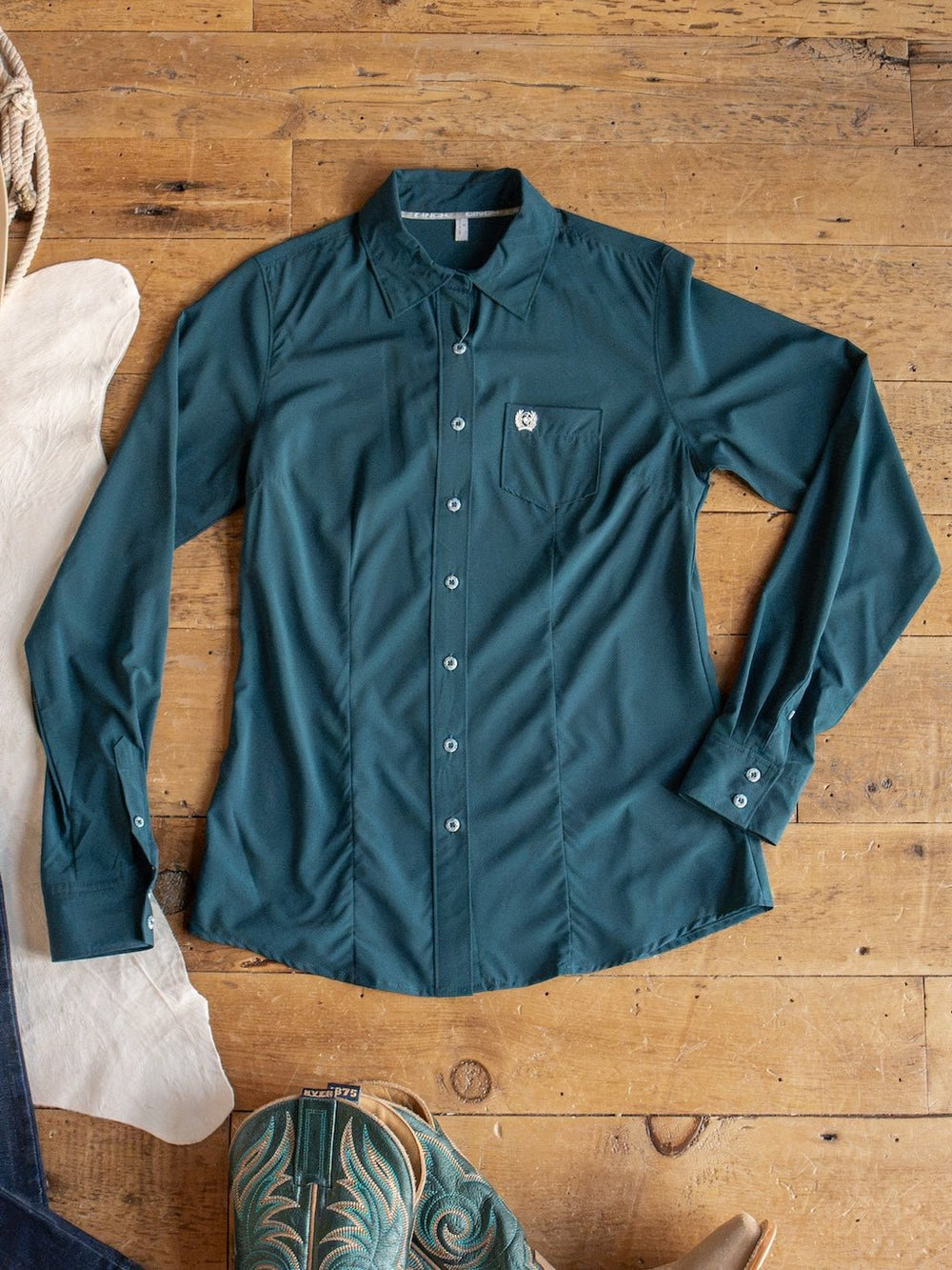 The Mankato Teal Mesh Performance Shirt by Savannah Sevens western life{&}style is laid flat on a wooden floor, featuring long sleeves, button-up front, UV defense, and a chest pocket. Cowboy boots and a white hat are partially visible near the shirt.