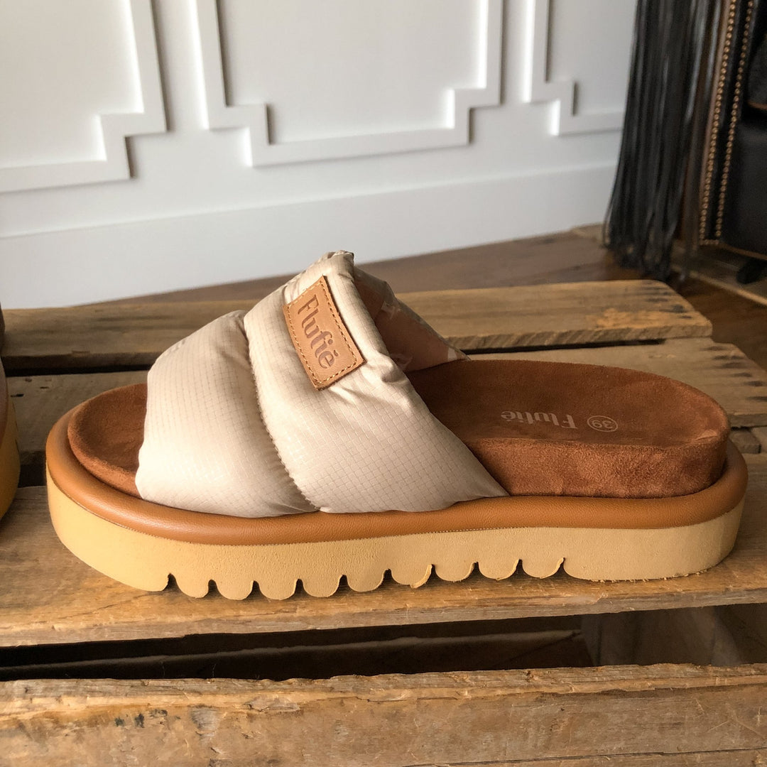 The Marfa {Natural} sandal by Savannah Sevens western life{&}style, in beige and brown with a cushioned strap, insole, and chunky shock-absorbing outsole, is displayed on a wooden surface against a white paneled wall.