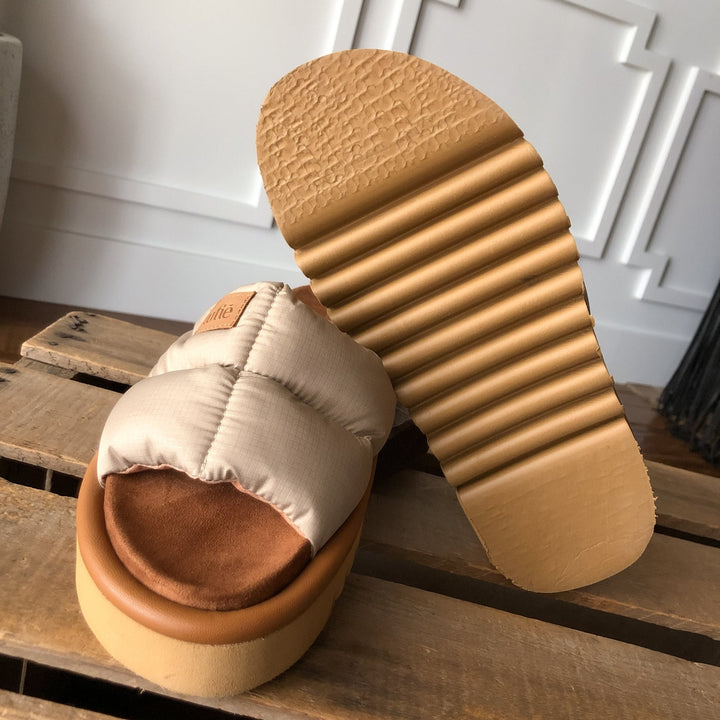 The Marfa {Natural} by Savannah Sevens western life{&}style features beige puffer sandals with cushioned insoles, shock-absorbing ribbed tan soles, a quilted top, and brown footbeds, displayed on a wooden surface.