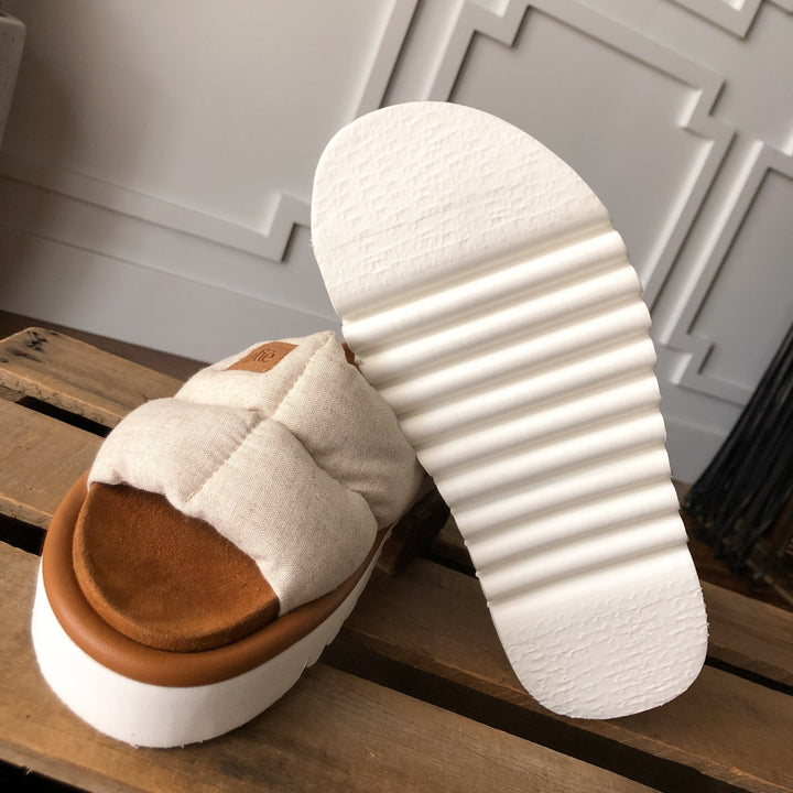 The Marfa {Oatmeal} puffer sandal by Savannah Sevens western life{&}style features a cushioned insole and thick, white shock-absorbing outsole, shown close-up on a wooden surface with a paneled wall behind.
