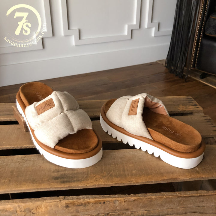 The Marfa {Oatmeal} puffer sandals by Savannah Sevens western life{&}style, featuring cushioned straps and a shock-absorbing outsole, are shown on a wooden surface indoors near a white wall. Sandals by Flufie. 