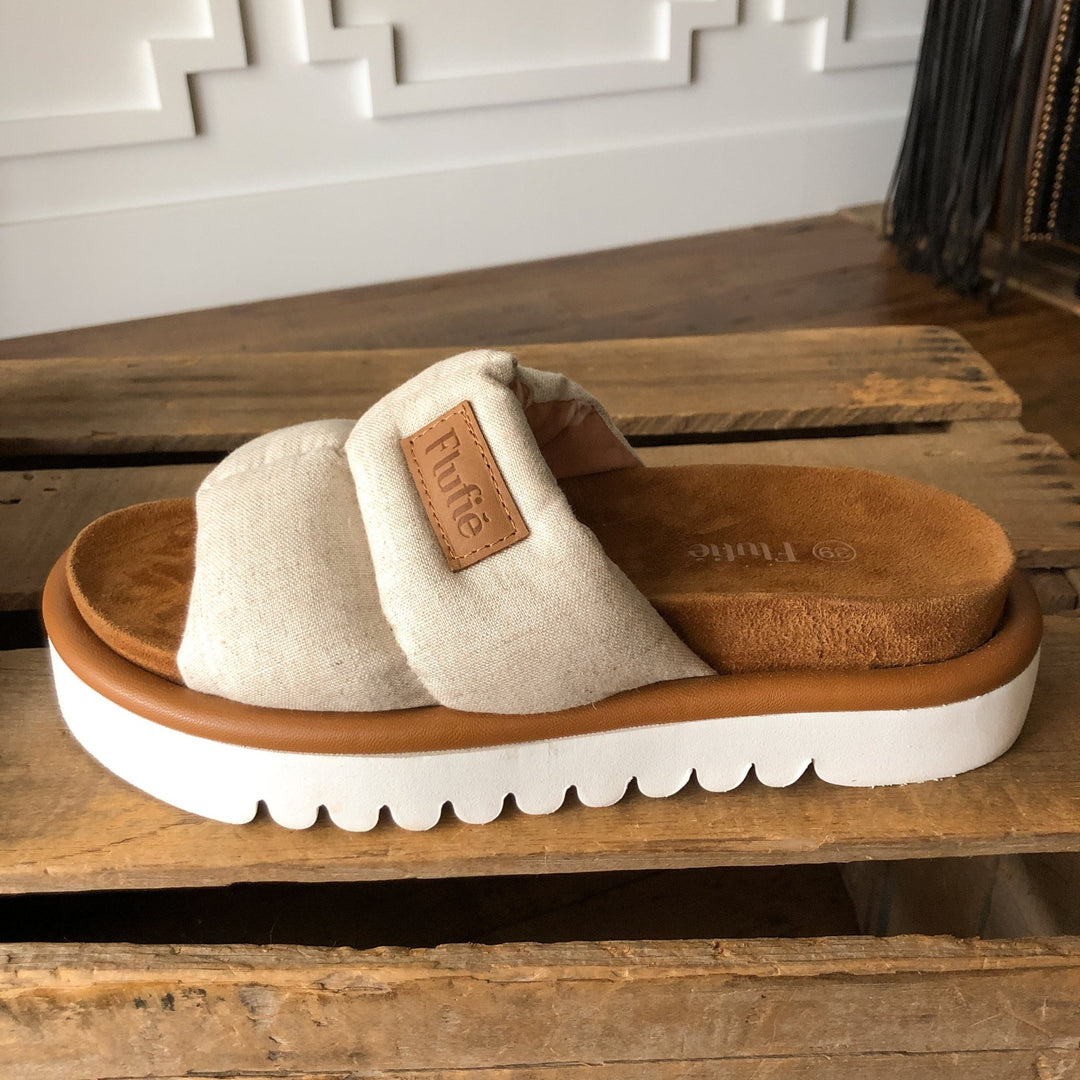 The Marfa {Oatmeal} by Savannah Sevens western life{&}style, a beige slip-on sandal with wide fabric strap, “Future” tan label, cushioned insole, and shock-absorbing outsole sits on a wooden surface indoors. Sandals by Flufie. 
