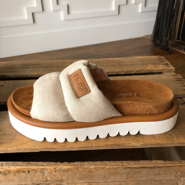The Marfa {Oatmeal} by Savannah Sevens western life{&}style, a beige slip-on sandal with wide fabric strap, “Future” tan label, cushioned insole, and shock-absorbing outsole sits on a wooden surface indoors. Sandals by Flufie. 