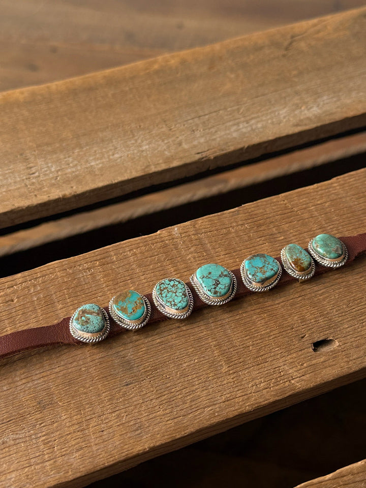 The Maricopa Leather Turquoise Bracelet by Savannah Sevens western life{&}style features seven Kingman turquoise stones, each set in sterling silver on a brown leather band, beautifully displayed on a rustic wooden surface.