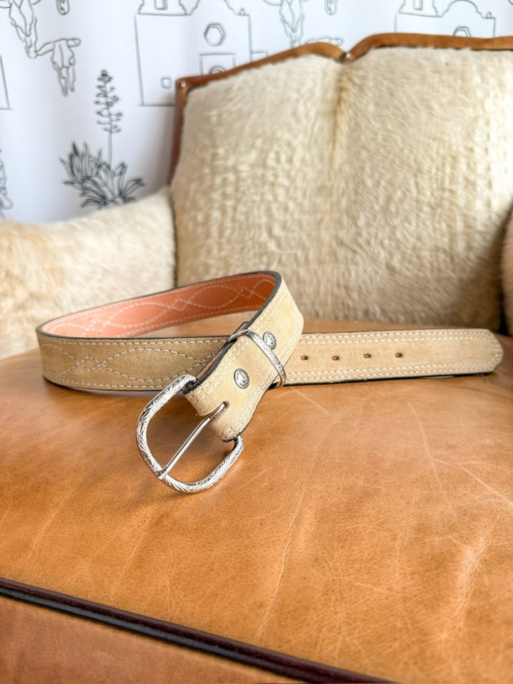 The Marietta Belt by Savannah Sevens western life{&}style, crafted in light tan rough out leather with a sterling silver buckle, sits on a tan leather cushion against a cream textured backrest and a wall with simple outlined drawings.