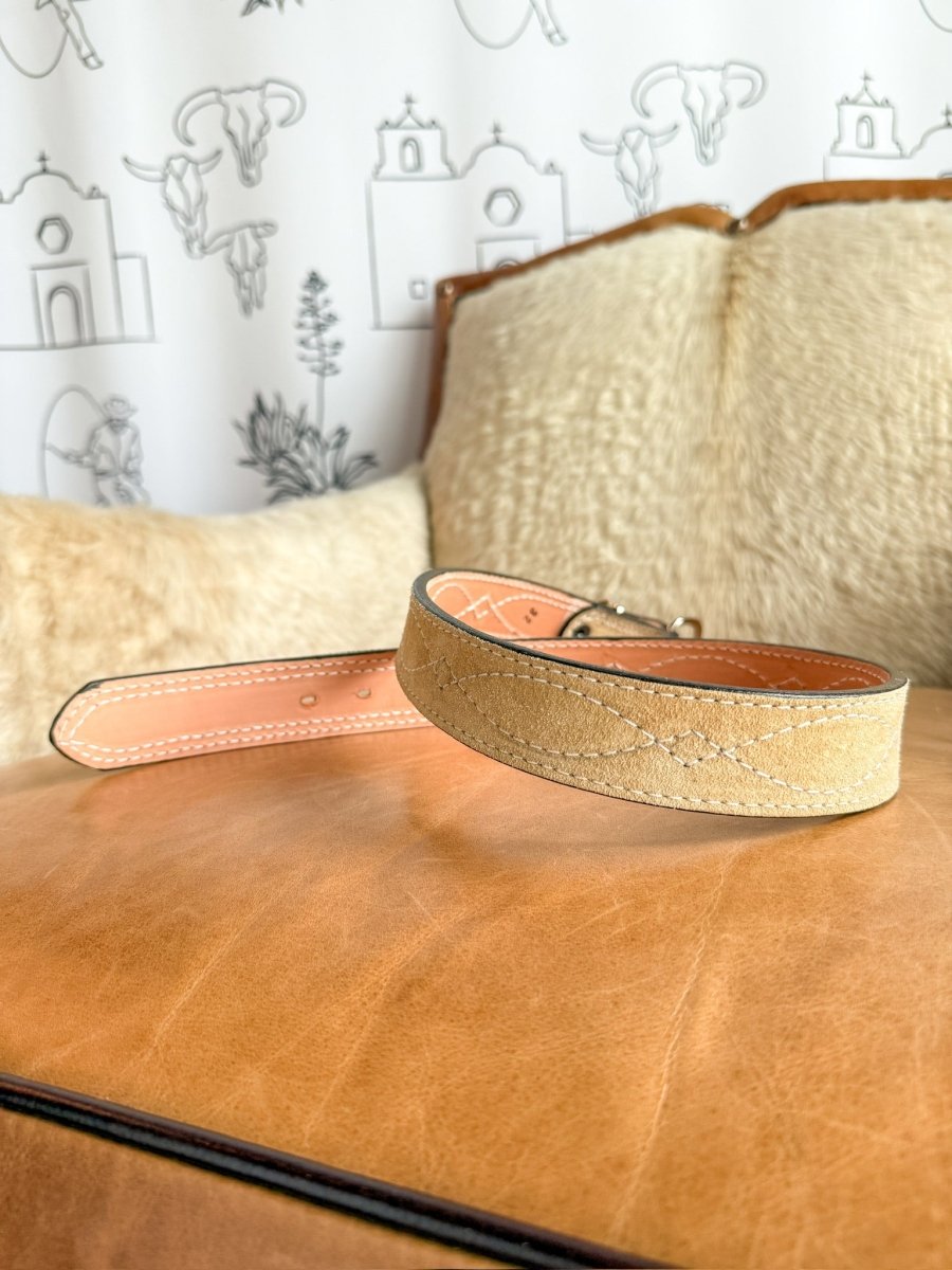 The Marietta Belt by Savannah Sevens western life{&}style, made of tan rough out leather with a suede texture, is shown on a brown leather cushion, with a beige pillow and southwestern-style line art on a white wall in the background.