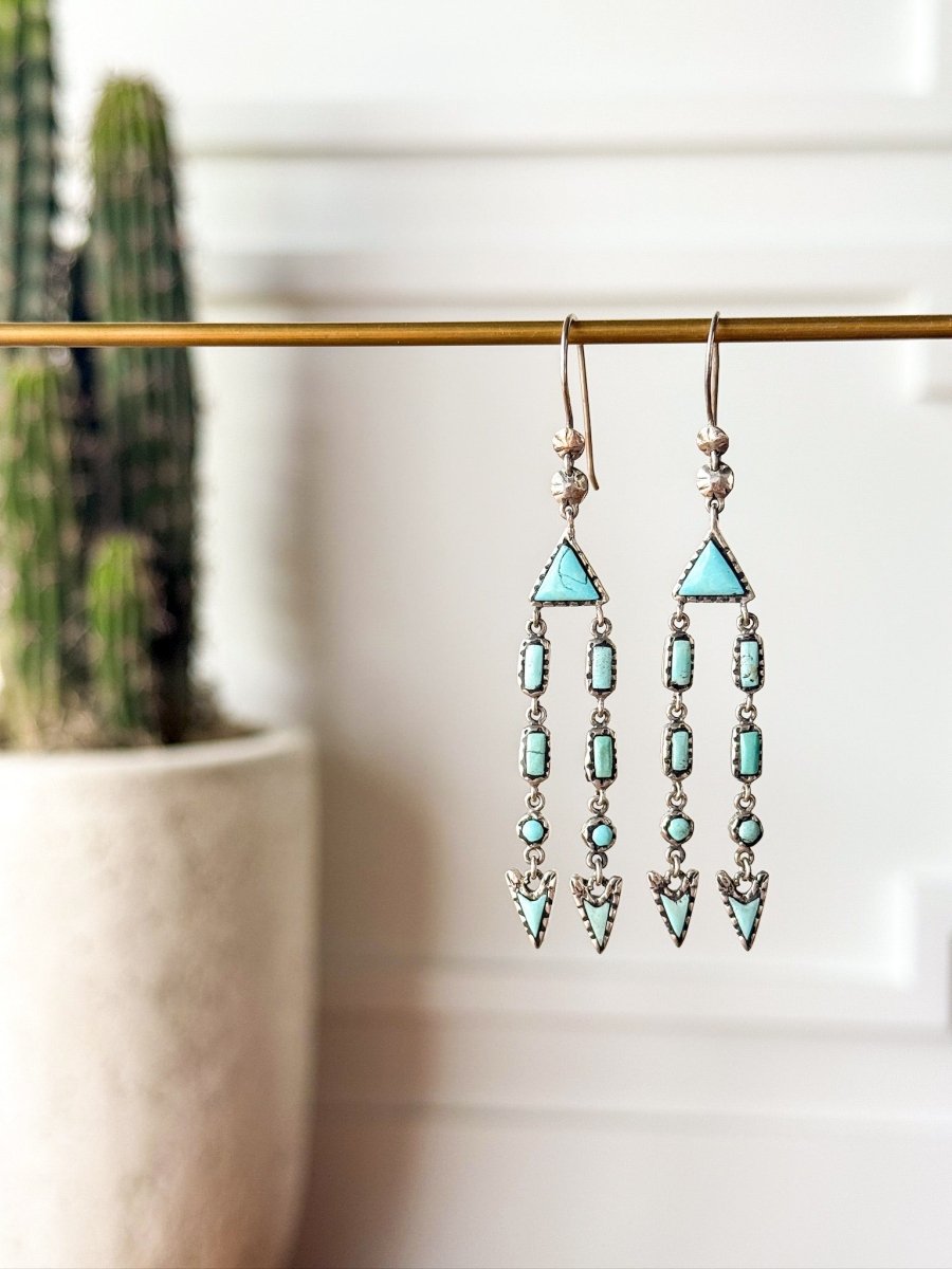 The Marilla Earrings by Savannah Sevens western life{&}style feature Kingman turquoise geometric drops hanging from a gold rod, with a blurred potted cactus in the background for a Southwest flair.