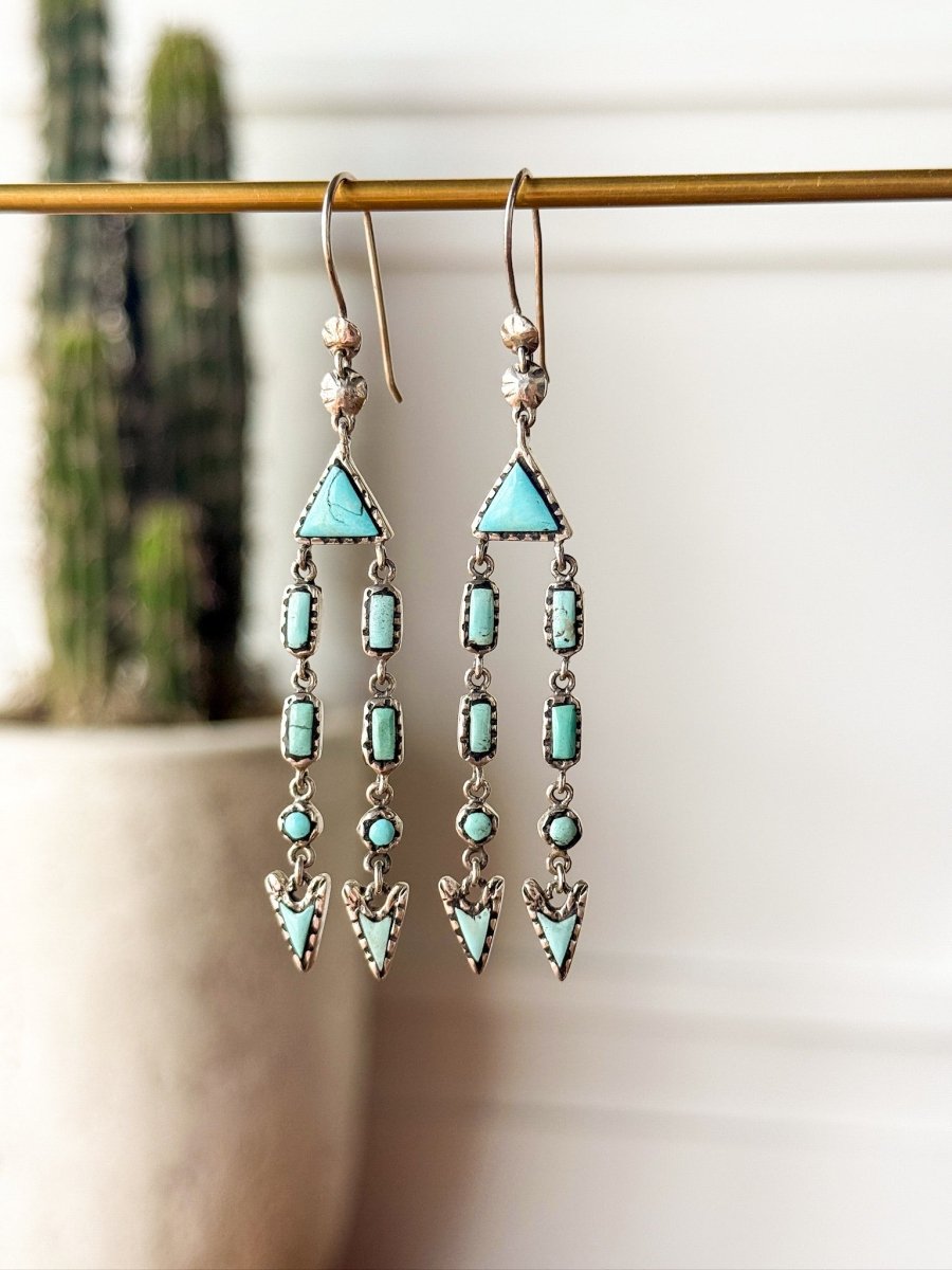The Marilla Earrings by Savannah Sevens western life{&}style feature long, geometric Kingman turquoise drops hanging from a brass rod, with a blurred cactus in the background evoking a southwestern jewelry vibe.