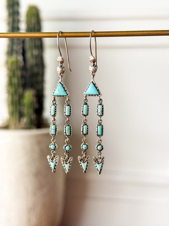 The Marilla Earrings by Savannah Sevens western life{&}style feature long, geometric Kingman turquoise drops hanging from a brass rod, with a blurred cactus in the background evoking a southwestern jewelry vibe.