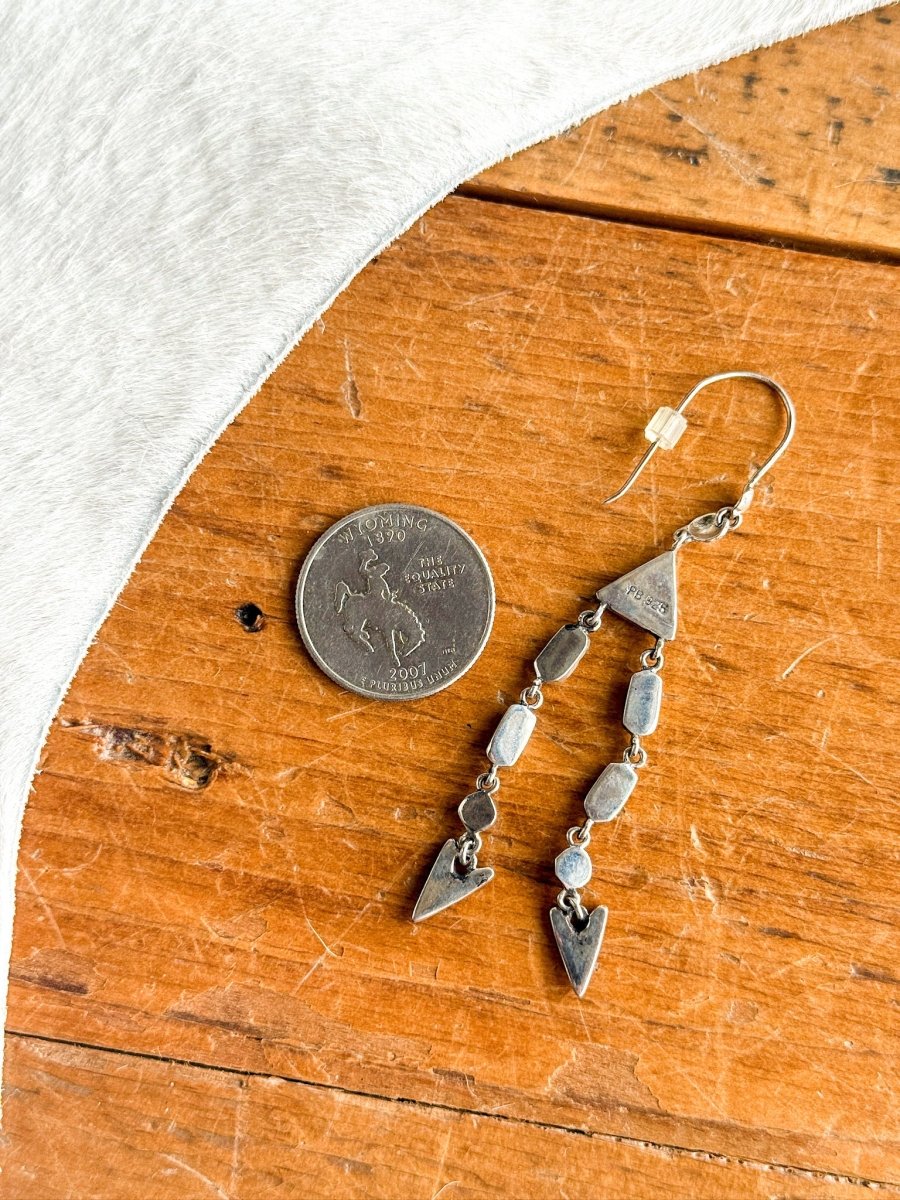 The Marilla Earrings by Savannah Sevens western life{&}style, featuring silver geometric dangles inspired by southwest jewelry, are shown beside a U.S. quarter on wood, with white fabric partially in view.