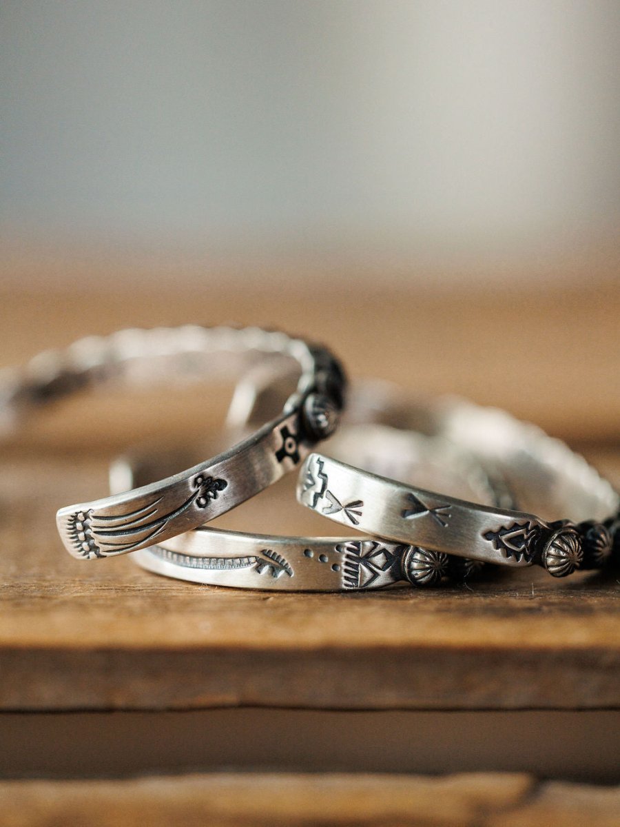 A close-up of several Marimar Cuff bracelets by Savannah Sevens western life{&}style, handmade in sterling silver with engraved patterns and symbols, stacked on wood against a softly blurred background.