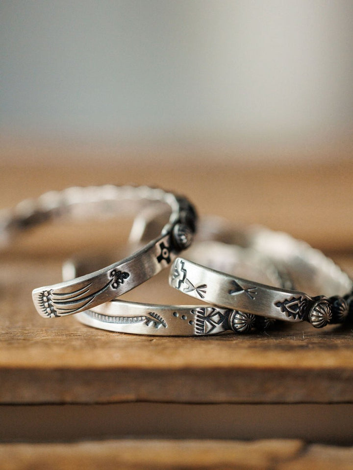 A close-up of several Marimar Cuff bracelets by Savannah Sevens western life{&}style, handmade in sterling silver with engraved patterns and symbols, stacked on wood against a softly blurred background.