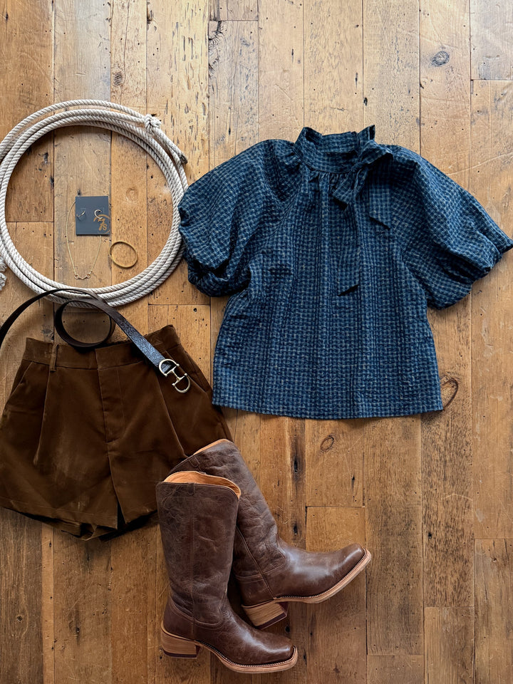 A navy Marion Plaid Puff Sleeve Top, brown shorts with a belt, and leather cowboy boots arranged on a wooden floor with jewelry.