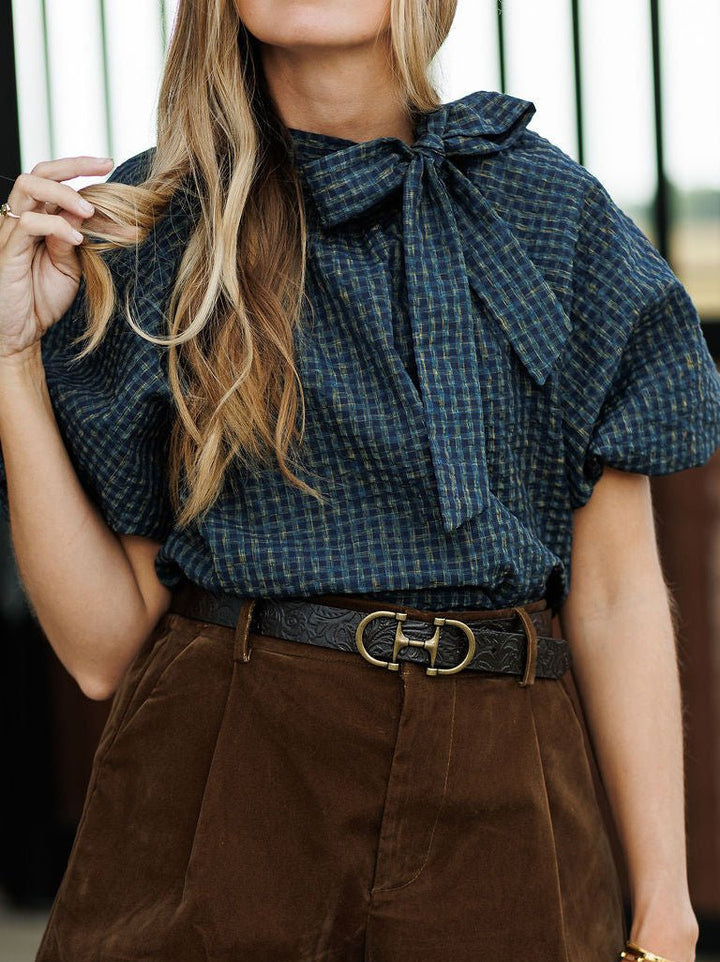 A woman wearing a navy plaid Marion Plaid Puff Sleeve Top with a bow, tucked into brown pants with a wide leather belt featuring a prominent gold buckle.