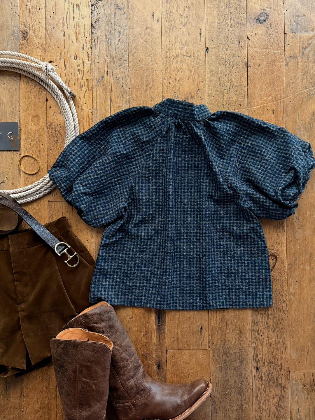 Marion Plaid Puff Sleeve Top by Savannah Sevens western life&style featuring a navy plaid cotton blend, high collar, and voluminous puff sleeves, arranged on a wooden floor with brown shorts, leather boots, and a coiled rope.
