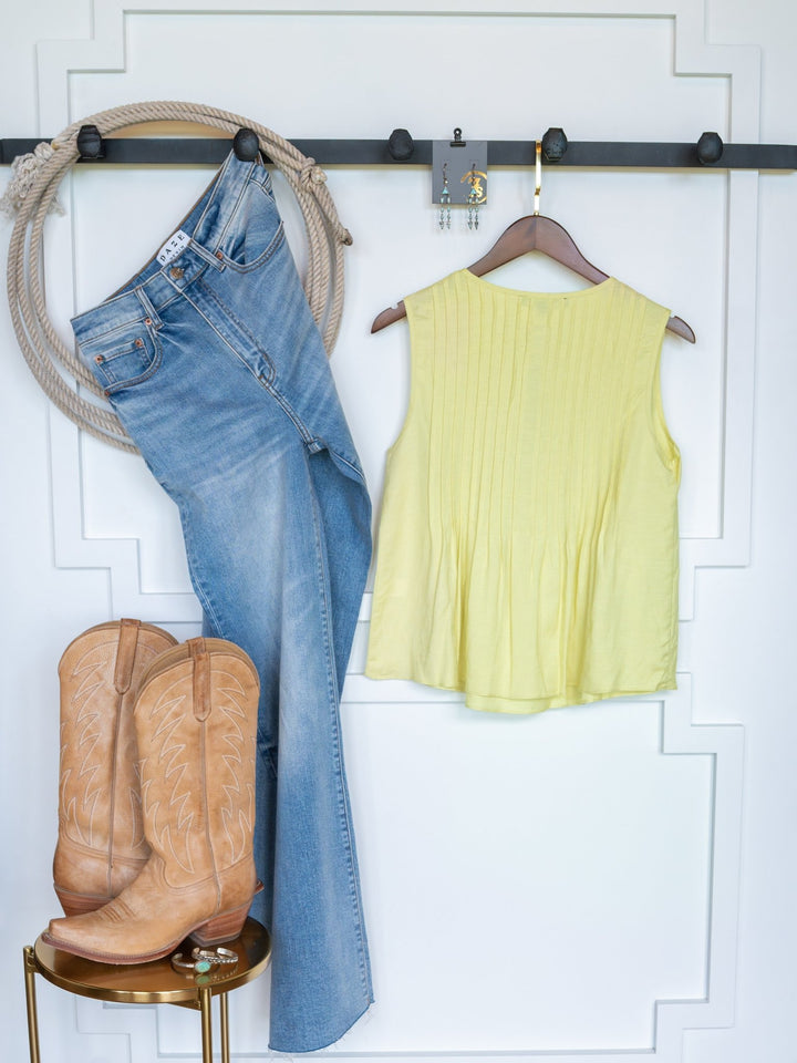 The Marlette jeans by Savannah Sevens western life{&}style hang with a linen blend sleeveless top on a wall rack, while tan cowboy boots sit on a round stool, creating a casual western-inspired look.