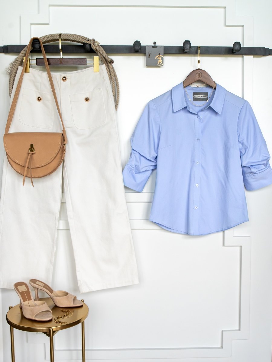 The Marlow Blue Tencel Button Up Top by Savannah Sevens western life{&}style hangs on a black wall rack beside white wide-leg pants and a tan shoulder bag, with beige high-heeled sandals on a small round gold table below.