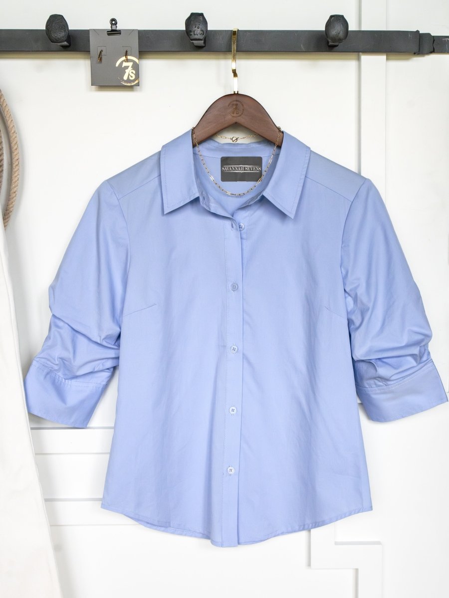 The Marlow Blue Tencel Button Up Top by Savannah Sevens western life{&}style, featuring ruched sleeves, hangs on a wooden hanger against a white wall and coat rack.