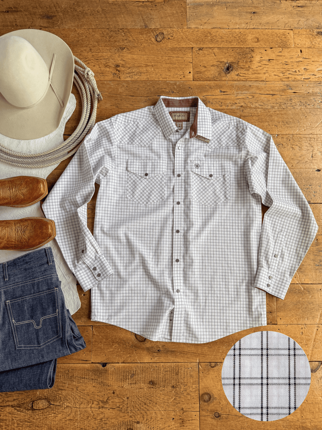 The Marshall Ariat Men’s Plaid Button Up from Savannah Sevens western life{&}style is neatly laid out on a wooden floor, surrounded by a cowboy hat, boots, and blue jeans. A close-up of the plaid pattern is displayed in the lower right corner.