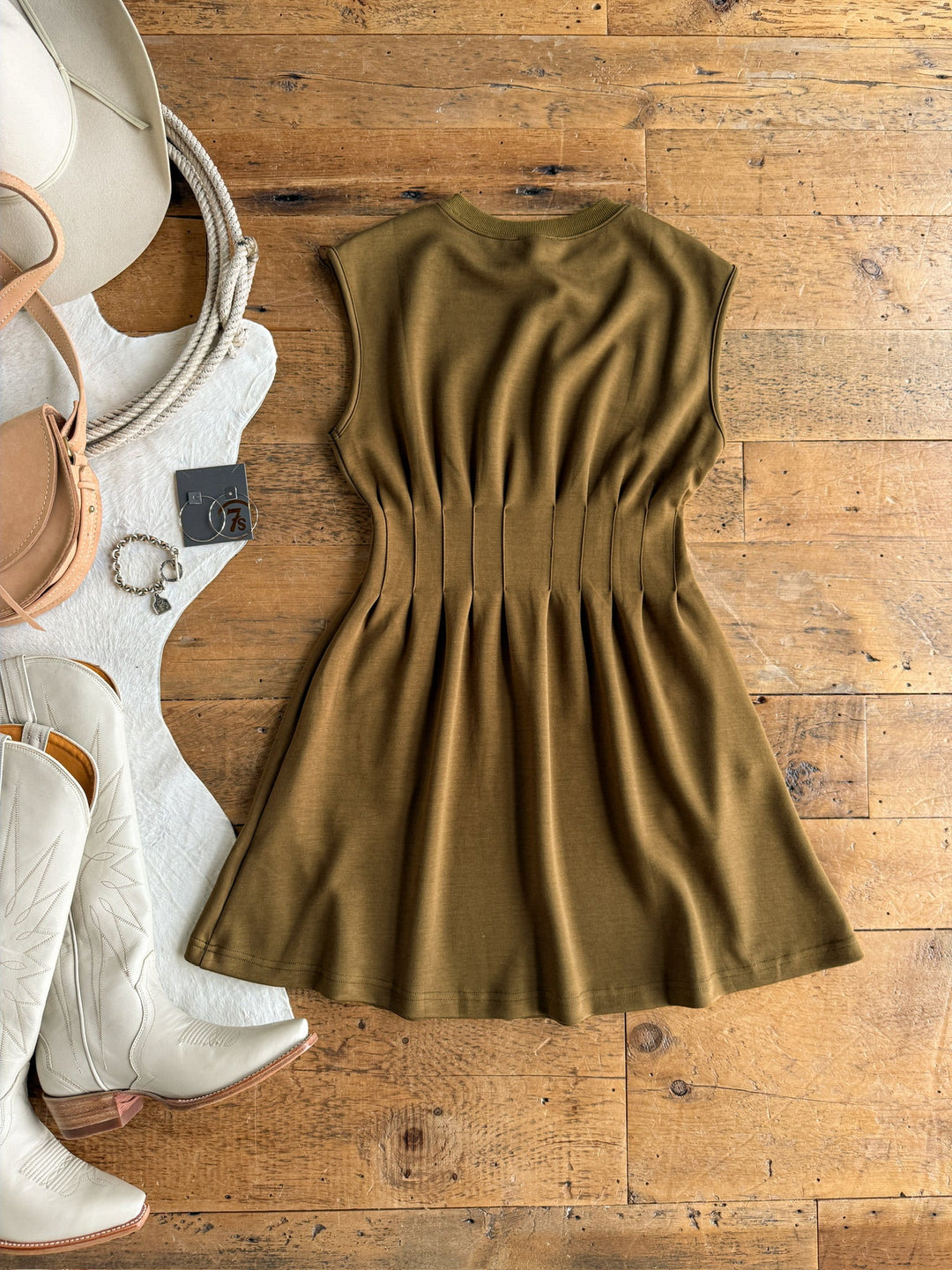 The Matewah Olive Pleat Waist Dress by Savannah Sevens western life{&}style is laid flat on a wooden floor, styled with cream cowboy boots, a tan handbag, and silver jewelry.