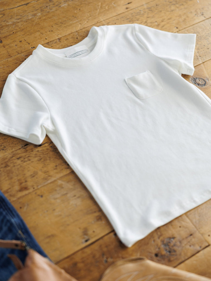 The Matteson White Basic Tee by Savannah Sevens western life{&}style, a classic crew neck with a chest pocket, is displayed on a wooden floor. Brown bag and blue jeans peek from the edges, showcasing its soft cotton blend fabric.