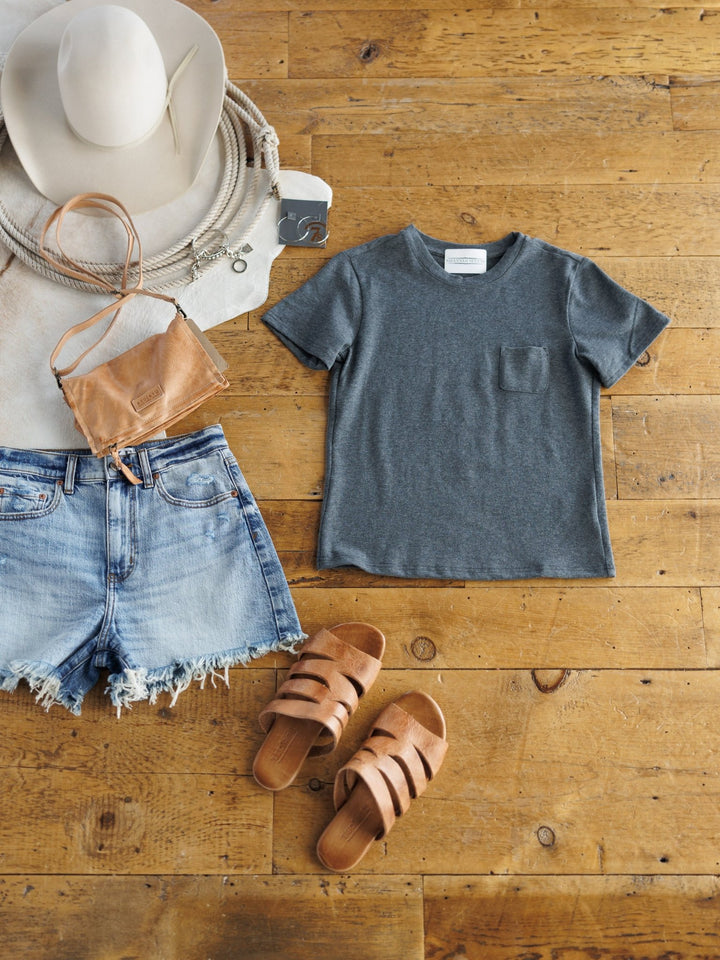 Flat lay of a summer outfit: light denim cutoff shorts, Matteson Charcoal Basic Tee by Savannah Sevens western life{&}style, tan leather sandals, a beige wide-brim hat, tan crossbody purse, and coiled rope on a wooden floor.