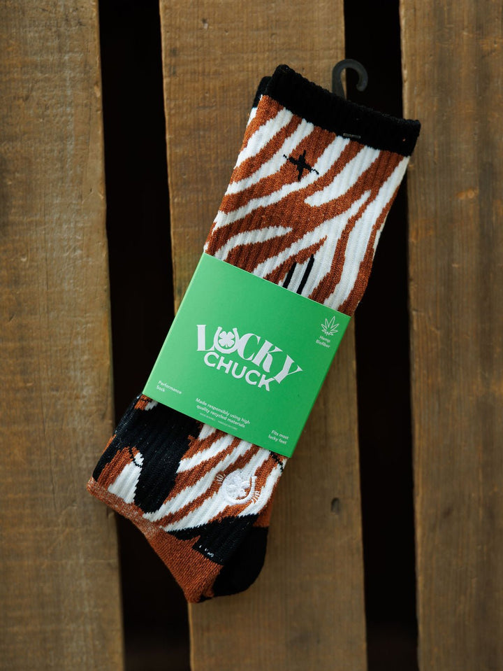A pair of Maverick Crew Socks by Savannah Sevens western life{&}style, featuring brown, black, and white zebra stripes in an odor-fighting hemp blend, wrapped in a green Lucky Chuck label and displayed on a wooden surface.