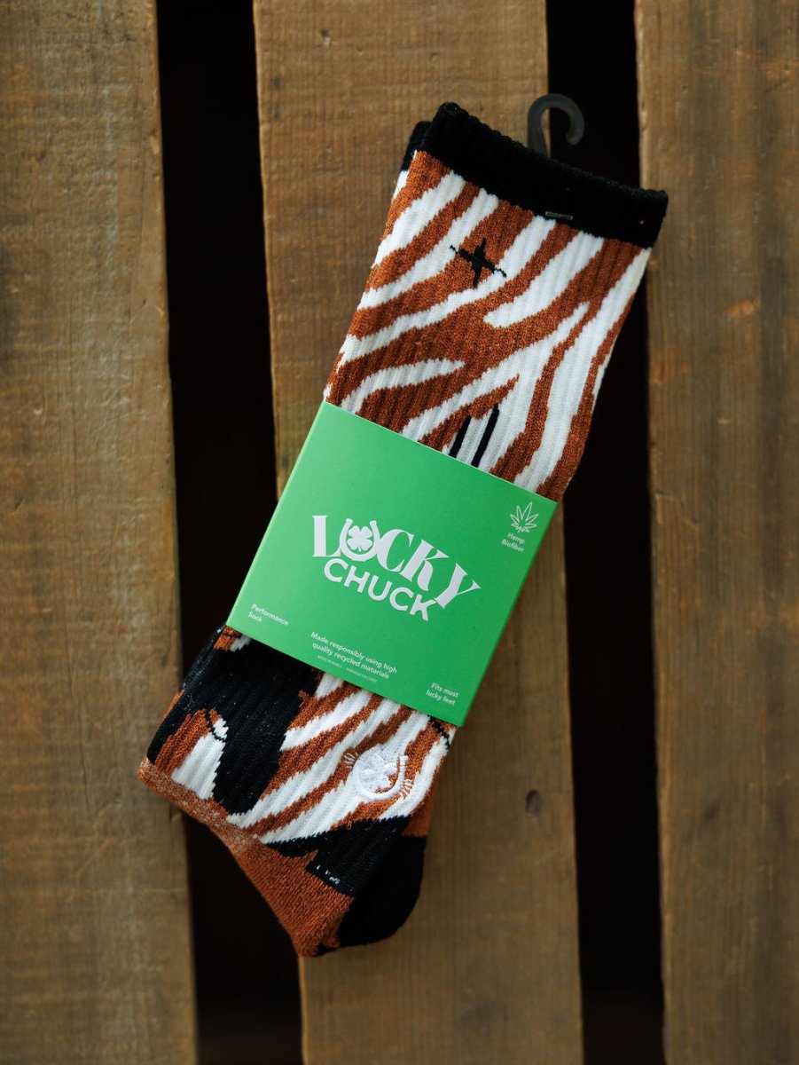 A pair of Maverick Crew Socks by Savannah Sevens western life{&}style, featuring brown, black, and white zebra stripes in an odor-fighting hemp blend, wrapped in a green Lucky Chuck label and displayed on a wooden surface.