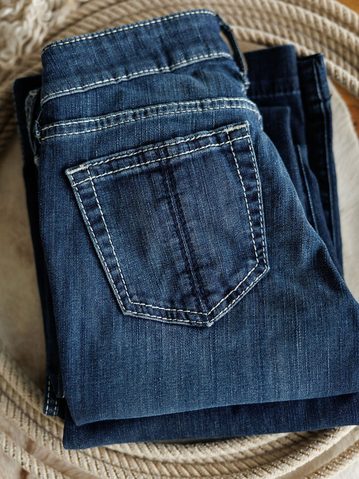 A folded pair of Ariat The Maverick Perfect Rise Trouser Jeans *Exclusive* in blue medium dark wash with white stitching rests on a coiled rope, showcasing the comfy no-gap waist, back pocket, and textured denim fabric.
