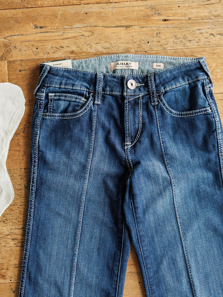 Ariat’s The Maverick Perfect Rise Trouser Jean *Exclusive* in medium dark wash denim features a comfy no-gap waist and white stitching, shown laid flat on a wooden surface with the Ariat label inside the waistband.