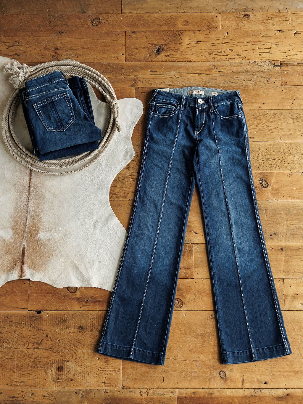 Two pairs of medium dark wash denim jeans rest on a wooden floor—one folded atop a coiled rope with a white cowhide rug, and the other is Ariat’s The Maverick Perfect Rise Trouser Jean *Exclusive*, showing off its flared legs and front seams.