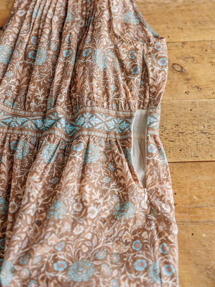 The Mayetta Cotton Floral Dress by Savannah Sevens western life{&}style features brown and blue floral patterns, a waistband detail, pleats, and a visible side pocket, shown close-up on a wooden surface.