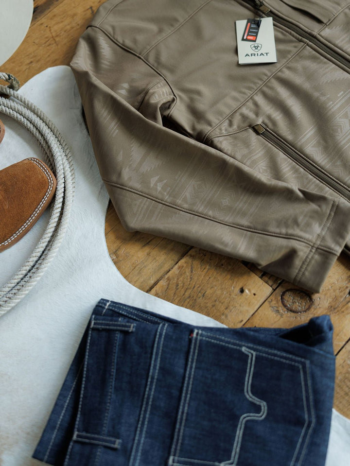 A Savannah Sevens western life{&}style McGregor Ariat Men's Southwest Insulated Jacket, blue jeans, a coiled rope, and a leather and white saddle blanket are displayed on wood. The wind- and water-resistant jacket elevates the Western vibe.