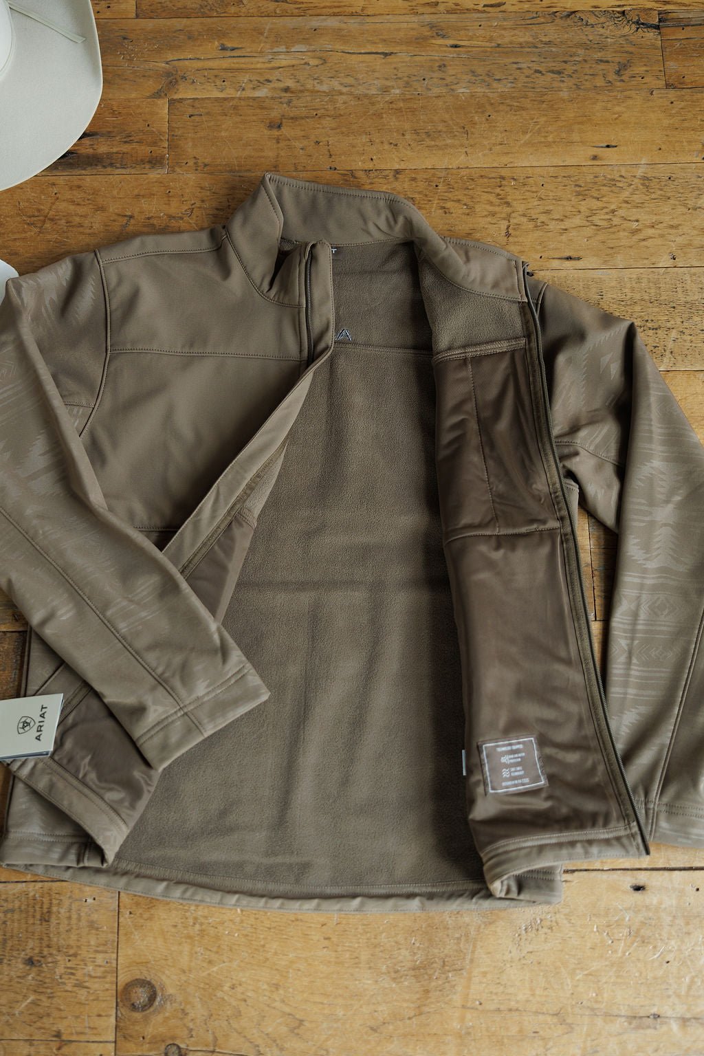 A McGregor Ariat Men's Southwest Insulated Jacket by Savannah Sevens western life{&}style lies open on a wooden floor, showing its lining and tags, with part of a white hat visible in the upper left corner.