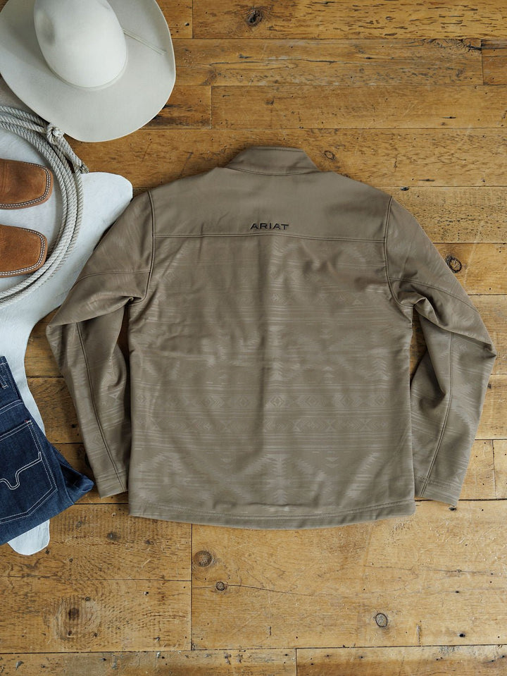 A Savannah Sevens western life{&}style McGregor Ariat Men's Southwest Insulated Jacket in tan lies flat on a wooden floor, paired with a white cowboy hat, coiled rope, brown boots, and blue jeans. The Ariat logo accents the back.