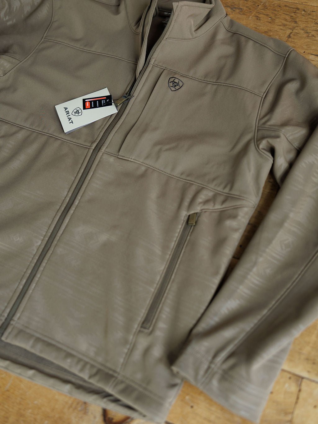 The McGregor Ariat Men's Southwest Insulated Jacket by Savannah Sevens western life{&}style is a tan soft shell with a chest pocket, small logo, and tag on the zipper pull. Wind and water resistant, it's ideal for versatile outdoor wear.