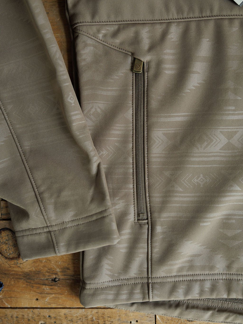 Close-up of the McGregor Ariat Men's Southwest Insulated Jacket by Savannah Sevens western life{&}style, showing its beige geometric-patterned sleeve, zippered pocket, soft shell insulation, and detailed stitching on a wooden surface.