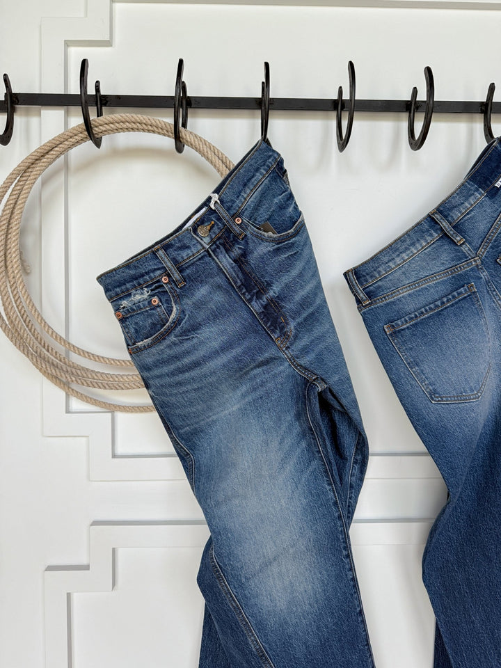 A pair of medium wash Savannah Sevens western life{&}style The McKinney High Rise Barrel Jeans hangs on a black metal hook rack mounted on a white wall, with a coil of rope and another pair of barrel jeans partially visible to the right.