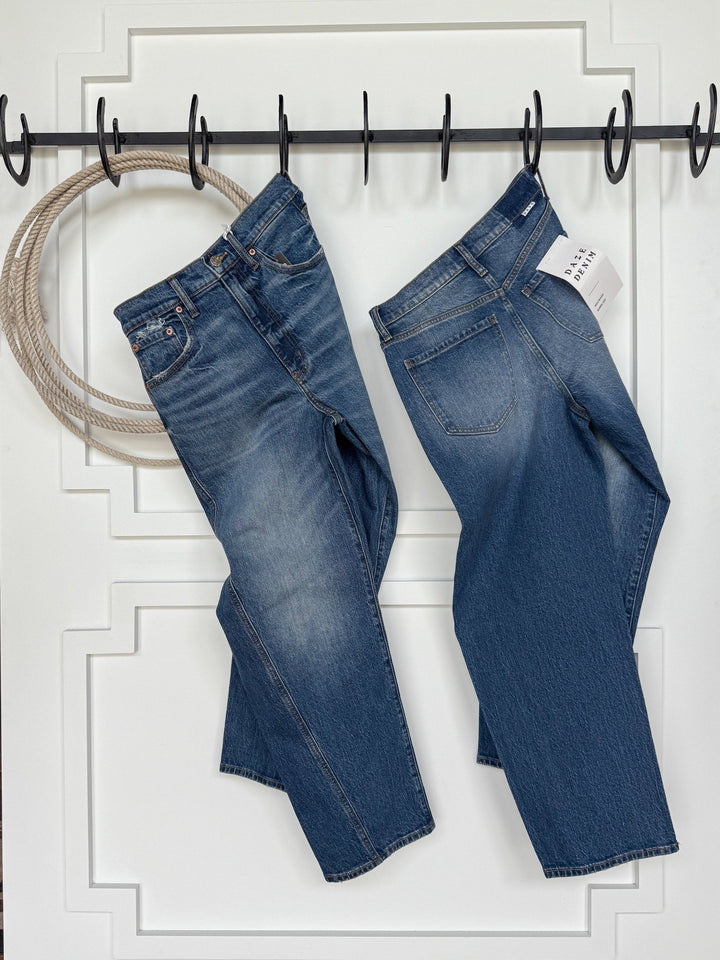 Two pairs of Savannah Sevens western life{&}style's The McKinney High Rise Barrel Jean hang by their belt loops on black hooks against a white paneled wall. A coiled rope hangs to the left, and a price tag is attached to the right pair.