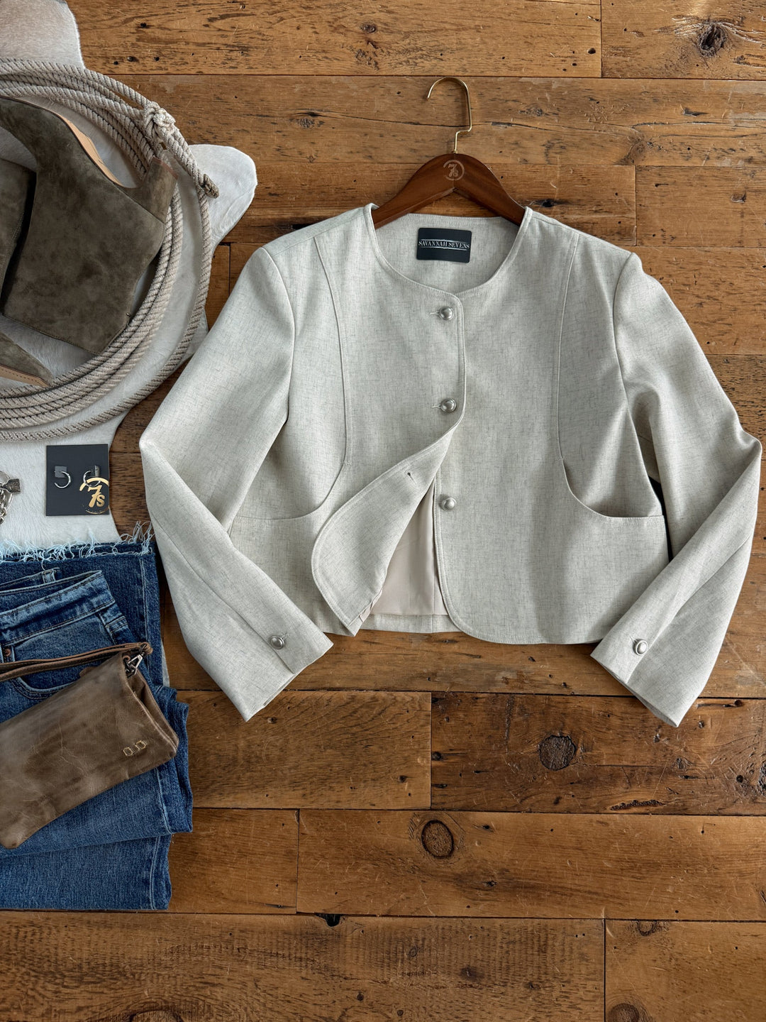 The Mead Oatmeal Tweed Jacket by Savannah Sevens western life{&}style, featuring a relaxed fit and cropped button-up design in light beige, is displayed on a wooden floor next to folded blue jeans, a taupe clutch, a gray rope-handle bag, and a small black card with a gold symbol.