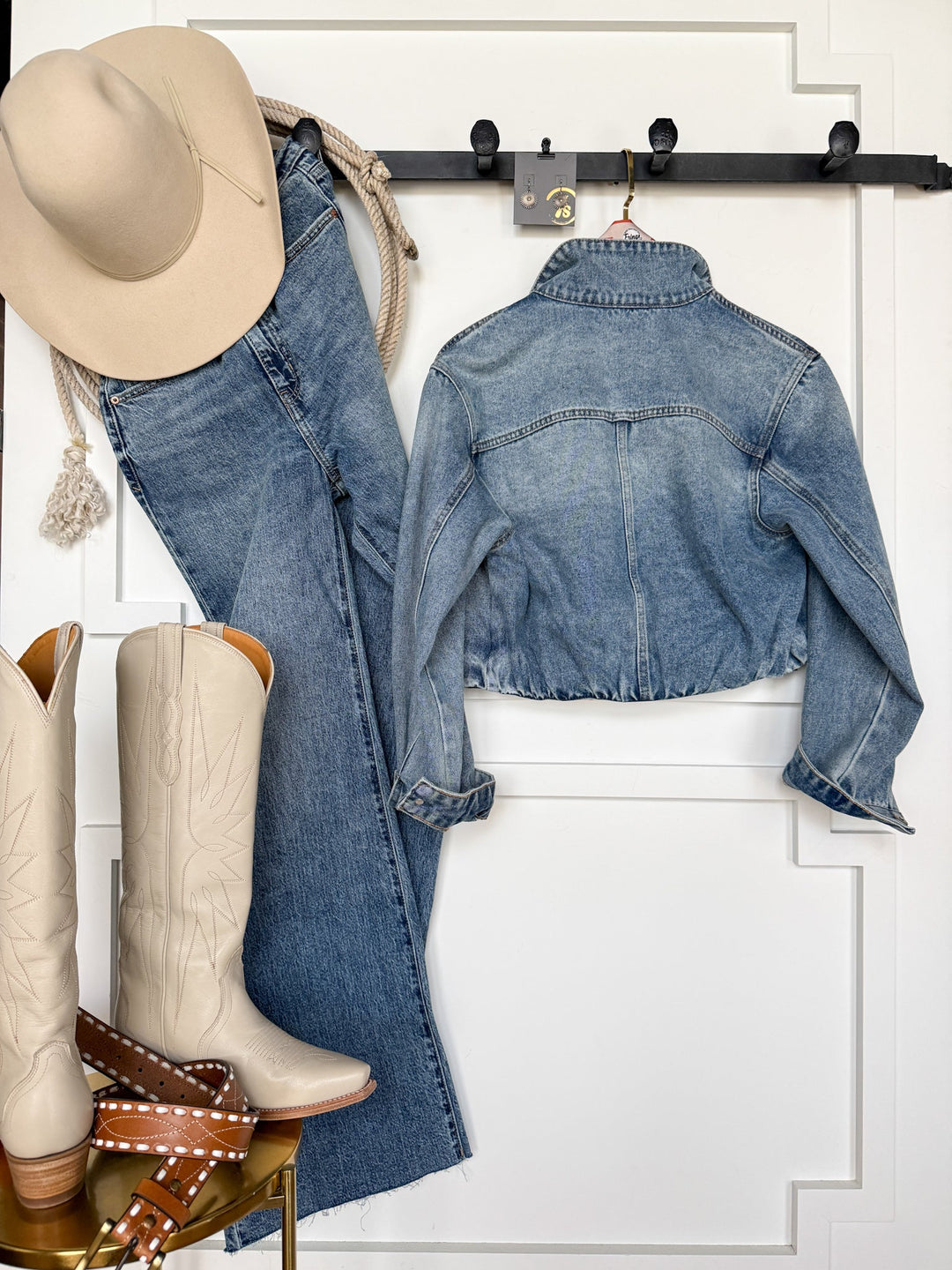 A beige cowboy hat, denim jeans, the Memphis Denim Jacket in a medium wash by Savannah Sevens western life{&}style, cowgirl boots, and a brown belt are arranged neatly against a white wall, evoking a western-inspired outfit.