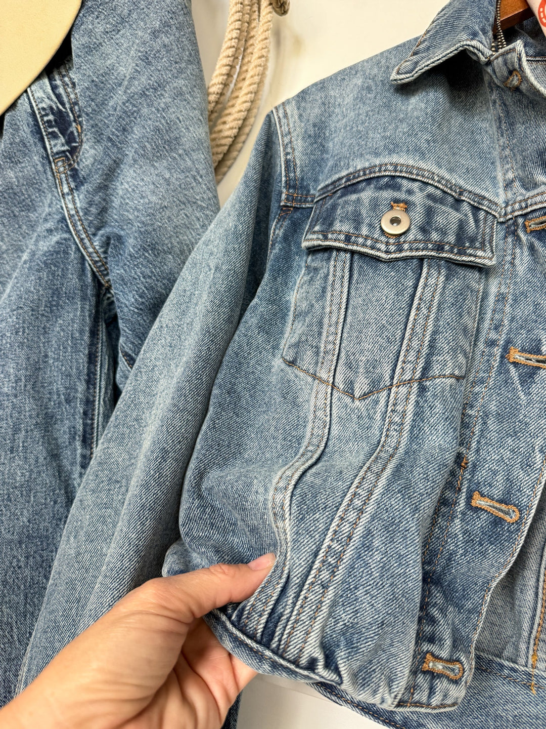 A hand holds the bottom of the Savannah Sevens western life{&}style Memphis Denim Jacket, featuring a medium wash, buttons, and chest pockets. Another Memphis Denim Jacket hangs in the background beside a thick beige rope.
