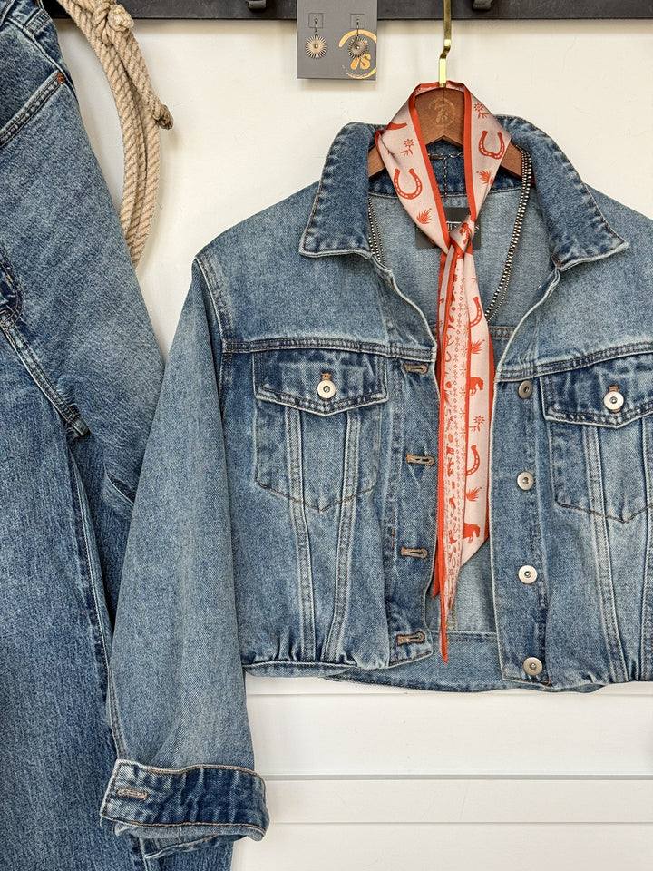 The Memphis Denim Jacket by Savannah Sevens western life{&}style, in a cropped medium wash, hangs on a hook with a pink patterned scarf tied at the collar. Blue jeans are draped beside it.